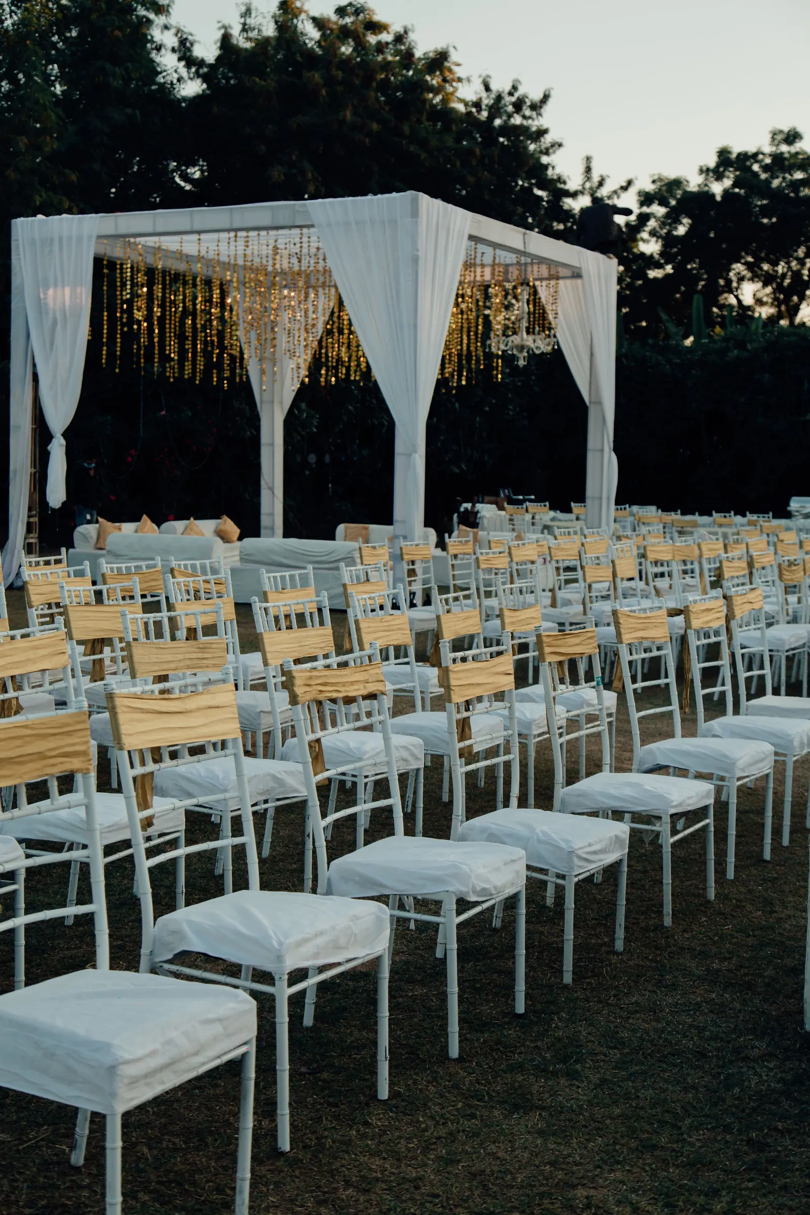 Outdoor wedding ceremony at dusk, with white chairs on a lawn facing a floral arch