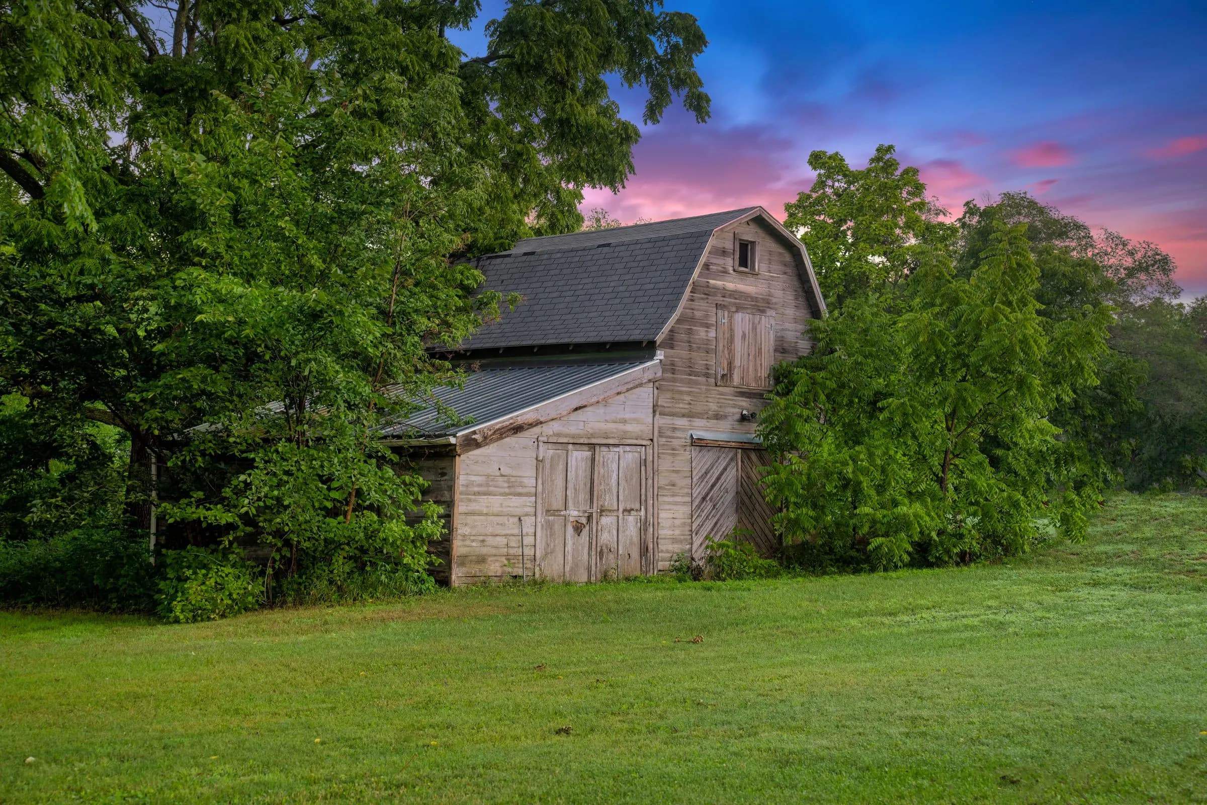 Wooden barn at golden hour across a green field, with trees behind it